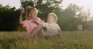 Little blonde girl with ponytail blows dandelion seeds ball to toddler brother face. Siblings sit and play on grass meadow in city park