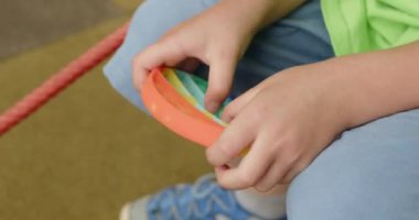 Boy wearing blue shorts and sneakers plays with rainbow pop it. Child fingers press small circles in toy sitting on yellow playground closeup