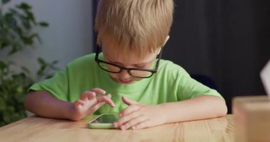 Preschooler boy wearing glasses enjoys playing games on smartphone. Concerned blond child makes funny drawings on device sitting at table