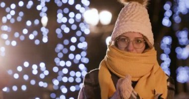 Young woman covers neck with orange scarf trying to get warm in cold evening. Lady stands on street against bright bokeh lights closeup