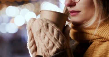 Blonde woman tries to get warm in winter drinking hot steaming tea in paper cup. Female person stands on street on blurred background closeup