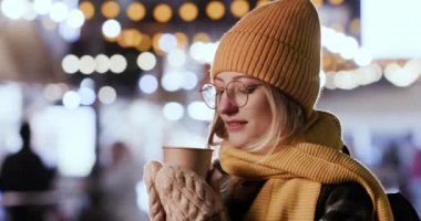 Blonde woman enjoys hot steaming tea in paper cup getting warm in cold winter evening. Happy lady stands against blurred Christmas lights closeup