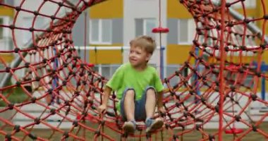 Preschooler boy plays alone jumping on red ropes attraction. Happy child excited being on kids playground against blurry yellow and grey buildings