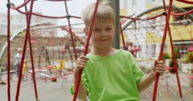 Happy preschooler boy enjoys swinging on red ropes on playground. Excited child plays on playground against yellow and grey buildings closeup