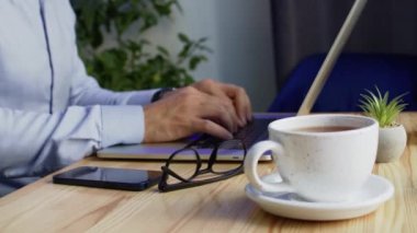 Freelancer works from home typing information on laptop. Cup of hot invigorating coffee stands on wooden table cooling down. Man puts on glasses