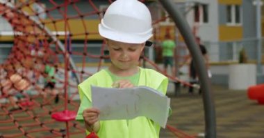 Preschooler boy enjoys playing builder on playground looking at papers. Excited child wearing special uniform and white helmet stands against yard