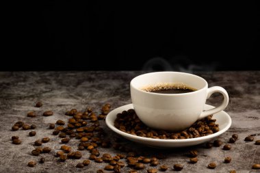 Beyaz bir fincanda sıcak kahvenin yakın çekimi beton zemine, kavrulmuş kahve çekirdekleri kahve fincanının içine konur ve fincandan duman ve aroma kokusu yükselir. Siyah, bulanık arkaplan