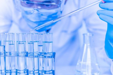 Koruyucu gözlük takan ve eldiven takan bir erkek bilim adamı aşıyı test etmek için kimyasallar damlatıyor. Biyokimyagerler yeni ilaçlar keşfetmek için deneyler yapıyorlar. Araştırmacılar bilim laboratuvarlarında çalışırlar..