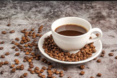 Beyaz bir fincandaki sıcak kahvenin yakın çekimi beton zemine konur, kahve fincanının içinde bir sürü kavrulmuş kahve çekirdeği bulunur ve fincandan dumanlar ve aroma kokusu yükselir. Bulanık arkaplan