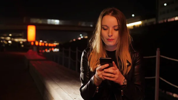 Kırmızı ışıklı bir sokakta oturan genç bir kadın akıllı telefon kullanıyor ve gülümsüyor.