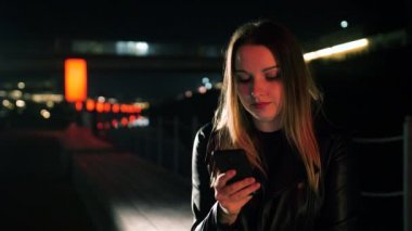 Genç bir kadın kırmızı ışıklı bir sokakta oturuyor ve akıllı bir telefona bakıyor.