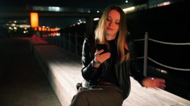 Kırmızı ışıklı bir sokakta oturan genç bir kadın akıllı telefon kullanıyor ve gülümsüyor.