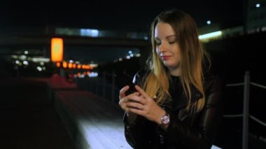 Kırmızı ışıklı bir sokakta oturan genç bir kadın akıllı telefon kullanıyor ve gülümsüyor.