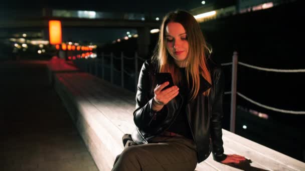 Jonge vrouw zit op een rood verlichte straat met behulp van een smartphone en glimlachen — Stockvideo