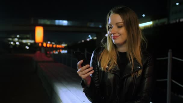 Junge Frau sitzt mit Smartphone auf einer rot beleuchteten Straße und lächelt — Stockvideo