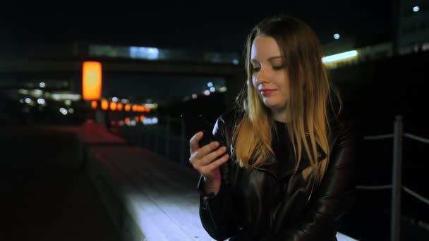 빨간 불이켜진 거리에 앉아 스마트폰을 사용하고 웃고 있는 젊은 여성 — 비디오