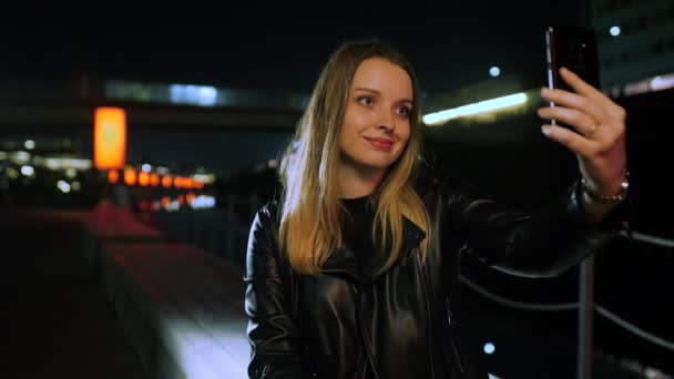 Kırmızı ışıklı bir sokakta oturan genç bir kadın akıllı telefondan selfie çekiyor. — Stok video