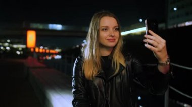 Kırmızı ışıklı bir sokakta oturan genç bir kadın akıllı telefondan selfie çekiyor.