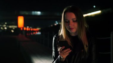 Kırmızı ışıklı bir sokakta oturan genç bir kadın akıllı telefon kullanıyor ve gülümsüyor.