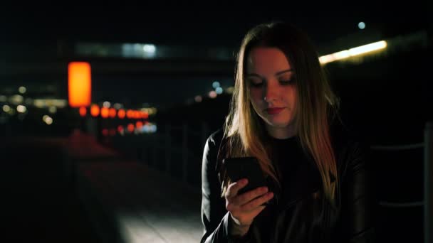 Kırmızı ışıklı bir sokakta oturan genç bir kadın akıllı telefon kullanıyor ve gülümsüyor. — Stok video