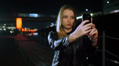 Kırmızı ışıklı bir sokakta oturan genç bir kadın akıllı telefondan selfie çekiyor.