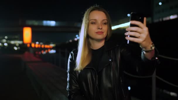 Mujer joven sentada en una calle iluminada por el rojo tomando una selfie en un smartphone — Vídeo de stock