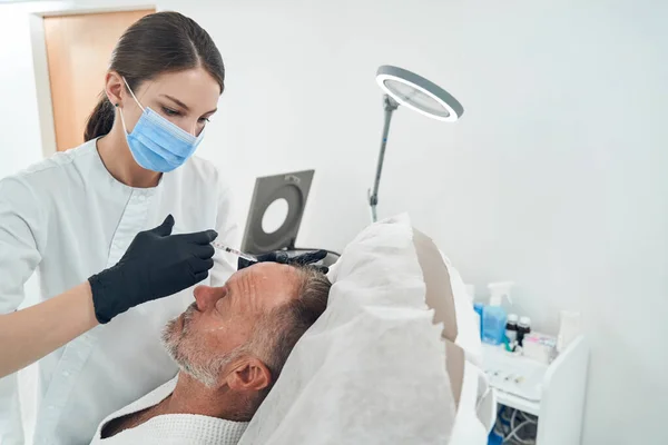 Kosmetolog i medicinsk mask gör injektion i människans panna — Stockfoto