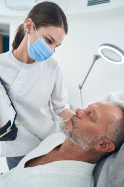 Güzellik uzmanı kozmetoloji kliniğindeki erkek müşteriye bakıyor.