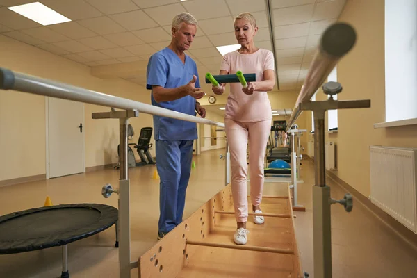 Fisioterapeuta que ayuda al paciente maduro a caminar entre barras paralelas en el hospital — Foto de Stock
