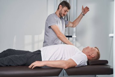 Fizyoterapist masaj masasında omuz yaralanması olan adamı kontrol ediyor.