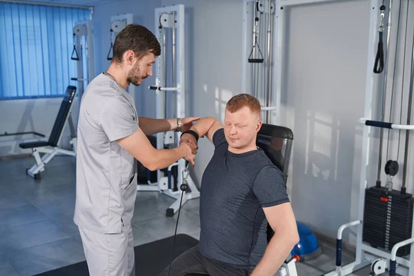 Doctor aids man with handicap lifting hand weight on rehab simulator — Stock Photo, Image