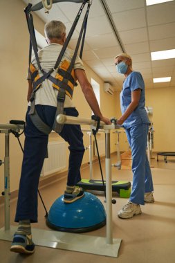 Rehabilitasyon odasındaki paralel barlarda yürüyen olgun bir adam.