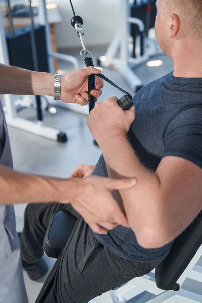 Hombre lesionado hace ejercicio de brazo en entrenador de fuerza con fisioterapeuta — Foto de Stock