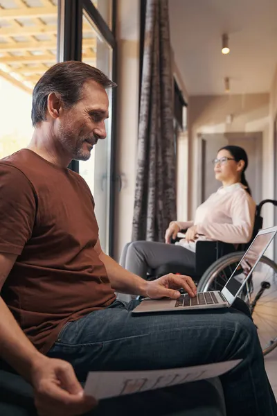 Χαμογελώντας άνθρωπος με φορητό υπολογιστή κοντά σύζυγος με αναπηρία σε εσωτερικούς χώρους — Φωτογραφία Αρχείου
