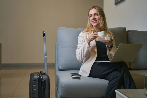 Mujer con café sonriendo a la cámara desde el sofá en el hotel — Foto de Stock
