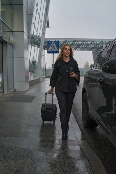 Frau in Mantel geht mit Koffer in der Nähe des Flughafens auf Straße — Stockfoto