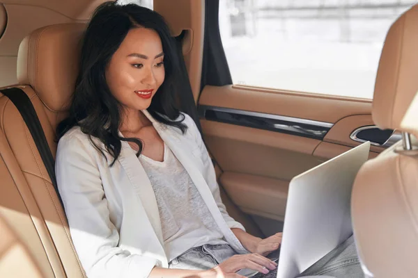 Positive business lady looking to the screen of her laptop in automobile — Stock Photo, Image