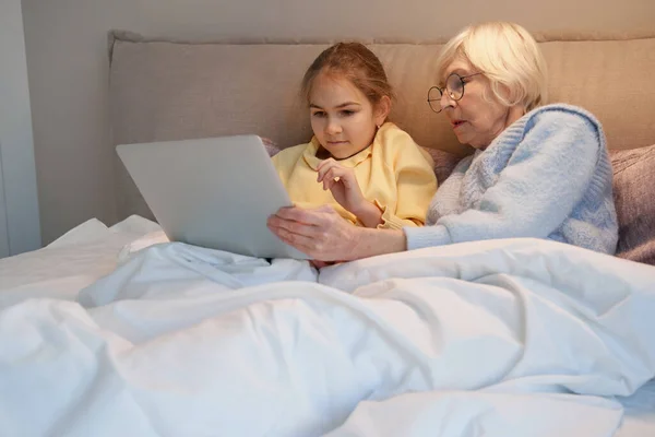 Милый ребенок и ее бабушка используют ноутбук в постели — стоковое фото