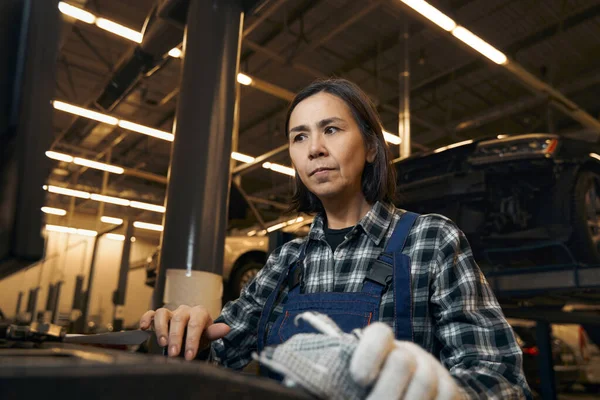 Serious brunette woman in mechanic uniform looking puzzled at workplace — Stockfoto