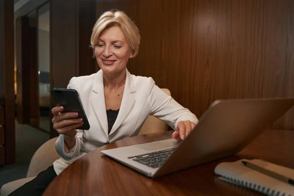 Χαμογελαστή επιχειρηματίας χρησιμοποιώντας φορητό υπολογιστή και smartphone στο δωμάτιο του ξενοδοχείου — Φωτογραφία Αρχείου