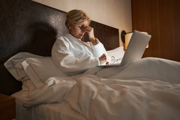 Mujer rubia cansada tocándose la frente mientras se queda en la cama — Foto de Stock