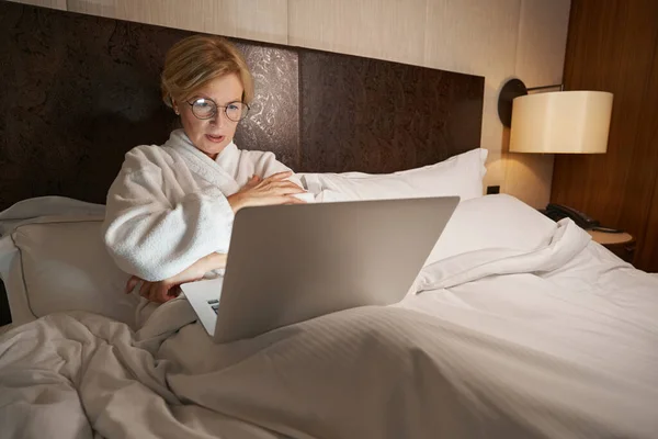 Geconcentreerde dame op zoek naar laptop tijdens het leggen in een gezellig bed — Stockfoto