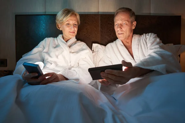 Caballero problemático y señora tranquila mirando los teléfonos inteligentes en la cama —  Fotos de Stock