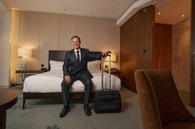 Otel yatağında otururken gülümseyen dost canlısı bir işadamı.