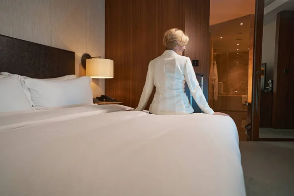 Фото блондинки в белом блейзере, сидящей на кровати отеля — стоковое фото