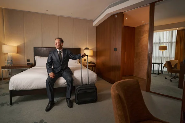 Энтузиастичный бизнесмен сидит на кровати и проверяет свой номер в отеле — стоковое фото