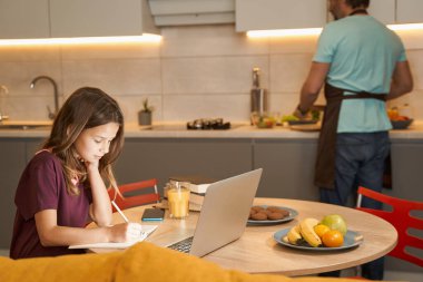 Mutfakta babasının yanında not defterine yazan bir ergen.