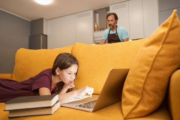 Дорослий чоловік готує на кухні, поки його дочка працює на ноутбуці — стокове фото