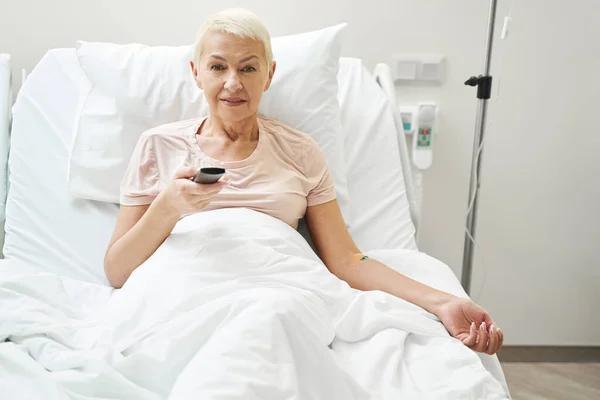Elderly patient clicking TV remote while under intravenous therapy — Foto de Stock