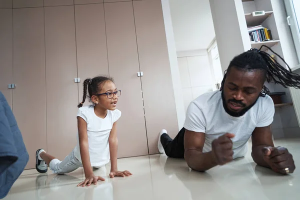 Adult african american father doing elbow plank next to daughter on floor — Stock Photo, Image